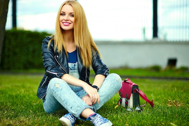 portrait of cute funny blond modern sexy urban young stylish smiling woman girl model in bright modern cloth outdoors sitting in the park on the green grass in jeans with pink bag