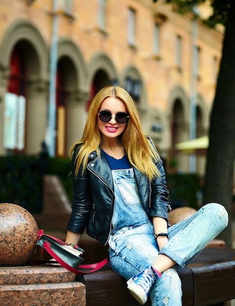 portrait of cute funny blond modern sexy urban young stylish smiling woman girl model in bright modern cloth outdoors sitting in the park in jeans on a bench in glasses