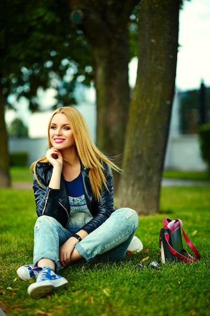 ピンクのバッグとジーンズの公園で屋外に座って明るいモダンな布でかわいい面白いブロンドモダンなセクシーな都市若いスタイリッシュな笑顔の女性女の子モデルの肖像画