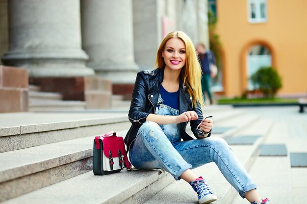 Портрет милые смешные современные сексуальные городские молодые стильные улыбающиеся женщина девушка модель в яркой современной ткани на улице, сидя в городе в джинсах с розовой сумкой