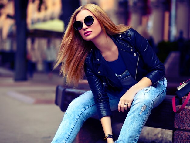 portrait of cute funny modern sexy urban young stylish smiling woman girl model in bright modern cloth outdoors sitting in the park in jeans on a bench in glasses