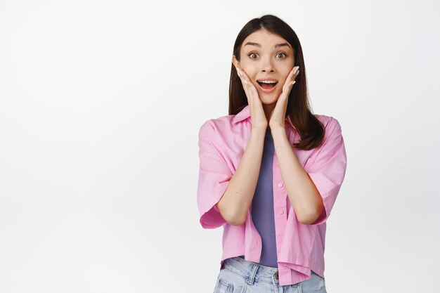 Portrait of cute girl gasp and look surprised saying wow looking with amazement at camera standing over white background