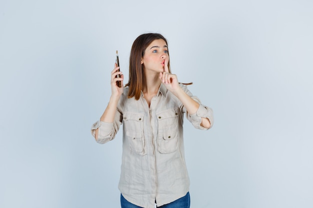 Portrait of cute girl holding mobile phone near ear, showing silence gesture in shirt and looking anxious