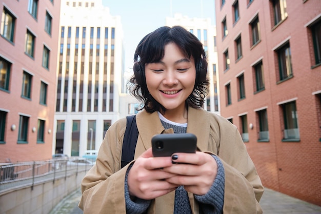 Portrait of cute girl tourist korean woman in headphones looks at mobile phone uses smartphone app m