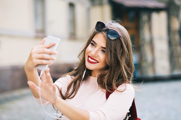 Free photo portrait of cute girl with long hair and vinous lips making selfie on street in city. she wears white shirt, smiling .