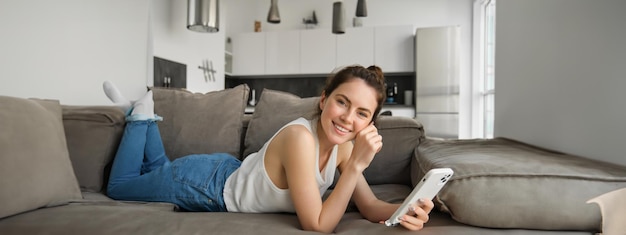 Foto gratuita ritratto di una giovane donna carina e felice sdraiata sul divano con in mano un telefono cellulare mentre usa uno smartphone