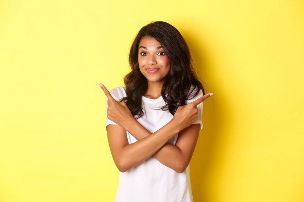 Portrait of cute indecisive african american girl pointing fingers sideways and shrugging asking adv...