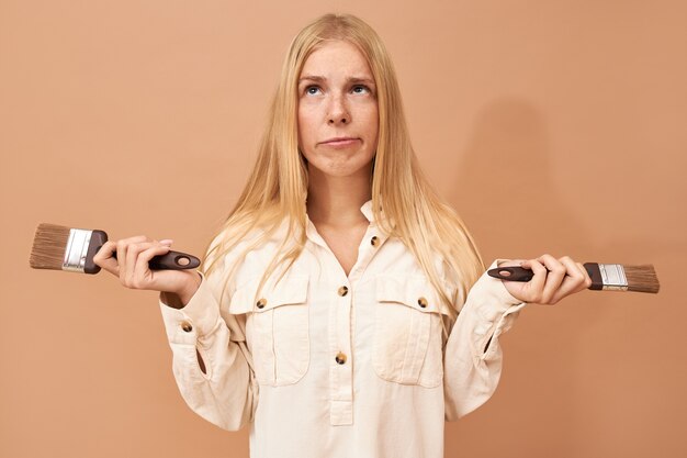 Portrait of cute indecisive young female with straight blond hair posing isolated with paint brushes, looking up with pensive facial expression, being doubtful