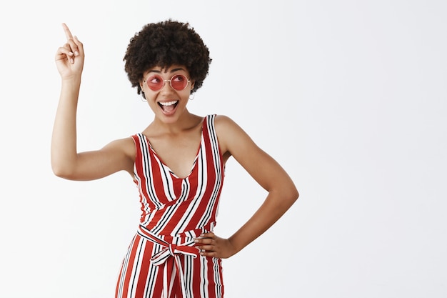 Portrait of cute joyful girl in striped stylish overalls and round sunglasses raising index finger, pointing up and making disco moved smiling broadly and gazing right