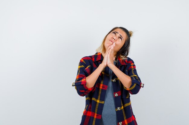 Free photo portrait of cute lady showing clasped hands in pleading gesture, looking up in checked shirt and looking sad front view