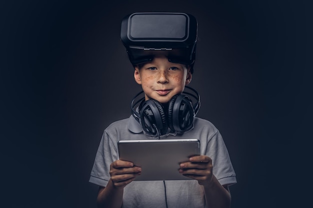 Free photo portrait of a cute little boy dressed in a white t-shirt, with virtual reality glasses and headphones is holding a digital tablet at a studio, looking at a camera, isolated on a dark background. educa