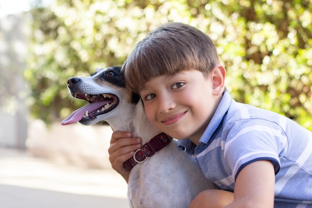 犬を抱き締めるかわいい男の子の肖像画。夏の日にTシャツとショートパンツと白い子犬の男子生徒。ペット、愛、子供の頃のコンセプト