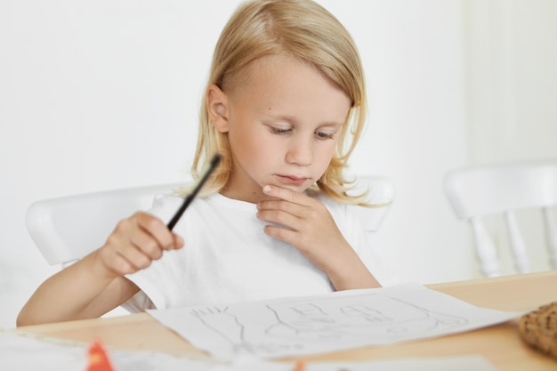 Портрет милого маленького мальчика со светлыми распущенными волосами, сидящего на стуле за деревянным столом, держа карандаш и трогательно подбородок, глядя на его рисунки. Ремесла, творчество, искусство, живопись и концепция детства