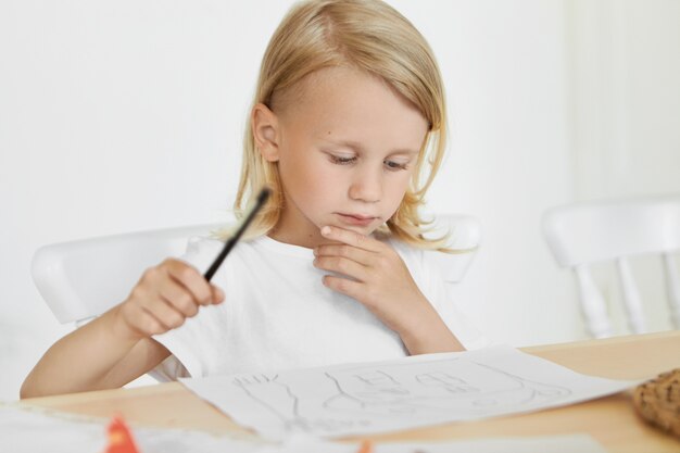 Portrait of cute little boy with blonde loose hair sitting on chair at wooden table, holding pencil and touching chin, looking at his drawings. Crafts, creativity, art, painting and childhood concept