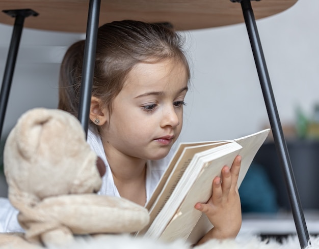 彼女のお気に入りのおもちゃで床に横たわって、家で本を読んでいるかわいい女の子の肖像画。
