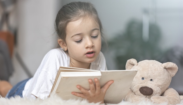 Portrait of a cute little girl reading a book at home, lying on the floor with her favorite toy.