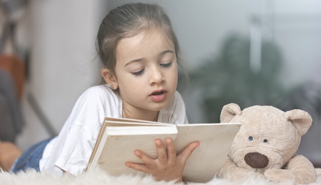 Портрет милой маленькой девочки, читающей книгу дома, лежа на полу со своей любимой игрушкой.
