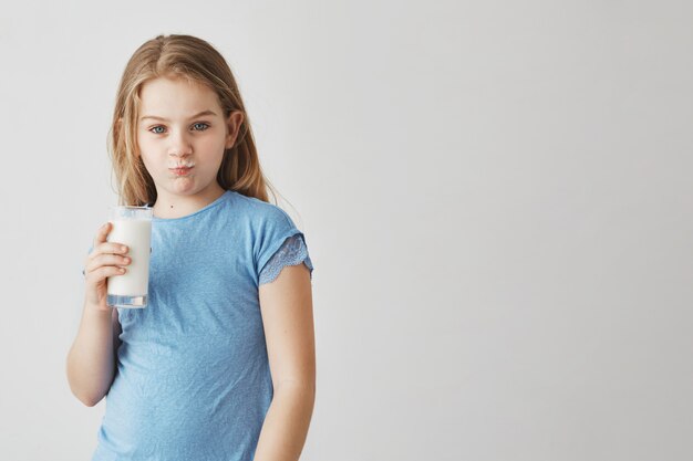 Portrait of cute little girl with blond long hair and blue eyes  with milk mustache and funny face expression, holding glass in hand.