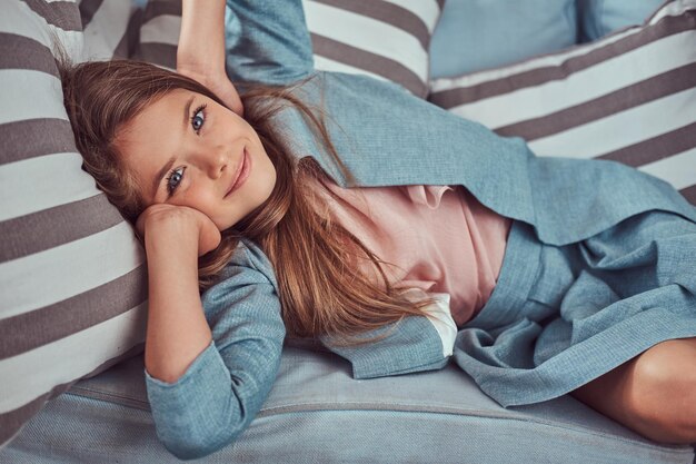Portrait of a cute little girl with long brown hair, piercing glance and charming smile, looking at a camera, lying on a sofa at home alone.