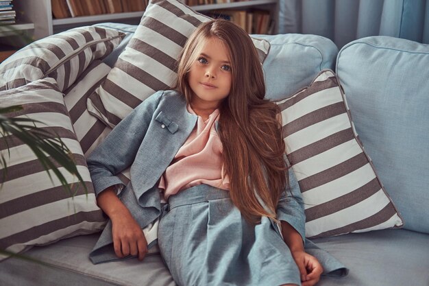 Портрет милой маленькой девочки с длинными каштановыми волосами и пронзительным взглядом, смотрящей в камеру, лежащей на диване дома одна.