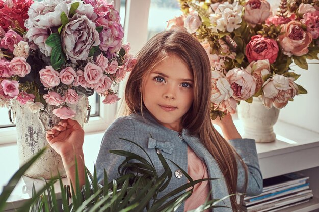 Portrait of a cute little girl with long brown hair and piercing glance wearing a stylish dress, posing with flowers against window at home.