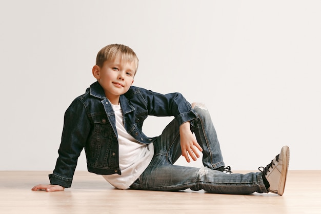 The portrait of cute little kid boy in stylish jeans clothes looking at camera against white studio wall.