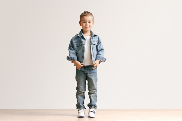 The portrait of cute little kid boy in stylish jeans clothes looking at camera against white studio wall.