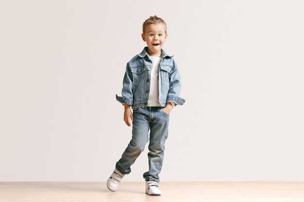 The portrait of cute little kid boy in stylish jeans clothes looking at camera against white studio wall.