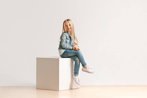 카메라를보고 웃 고, 흰색 스튜디오 벽에 앉아 세련 된 청바지 옷에 귀여운 꼬마 소녀의 초상화. 키즈 패션 컨셉