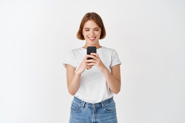 스마트폰 화면을 읽고 흰색 배경에 서서 휴대폰으로 수다를 떨며 웃고 있는 귀여운 자연 소녀의 초상화