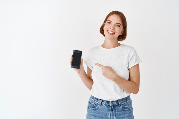 Portrait of cute natural girl smiling, pointing finger at smartphone screen. Woman showing app on display, wearing jeans with t-shirt, white wall