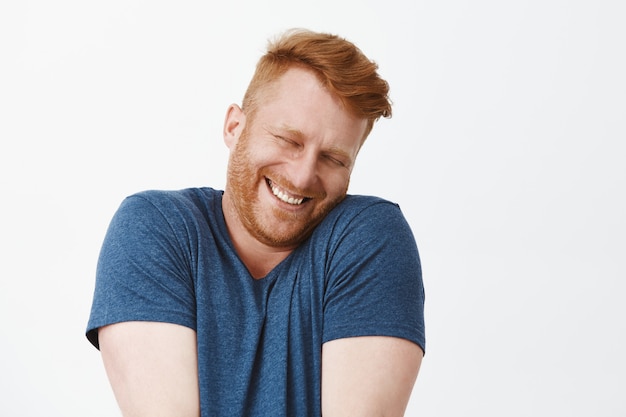 Free photo portrait of cute pleased and satisfied happy and funny redhead man in blue t-shirt, shrugging and leaning face on shoulder while smiling broadly, blushing from joy and happiness