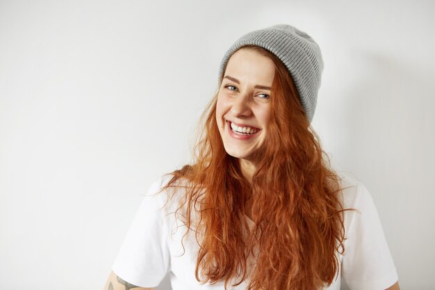 Портрет симпатичной рыжей девушки в серой зимней шапке и белой футболке улыбается