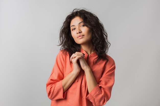 Portrait of cute shy beautiful woman in orange stylish shirt, curly hair, smiling, holding her hands together, isolated