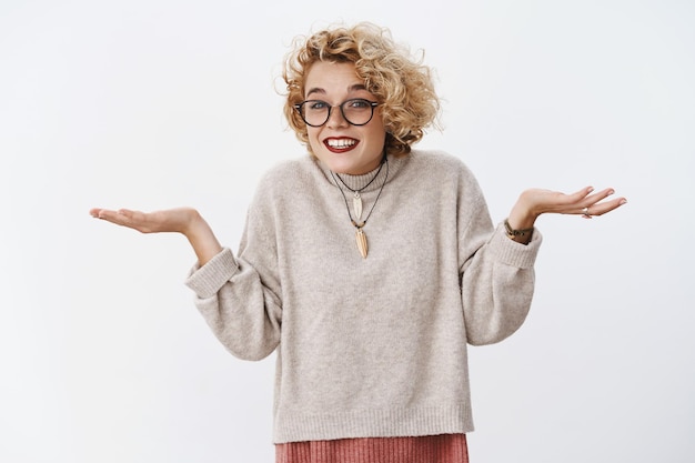 Portrait of cute and silly blond girl with short haircut and glasses shrugging and smiling with apology holding hands sideways in confused gesture over white wall