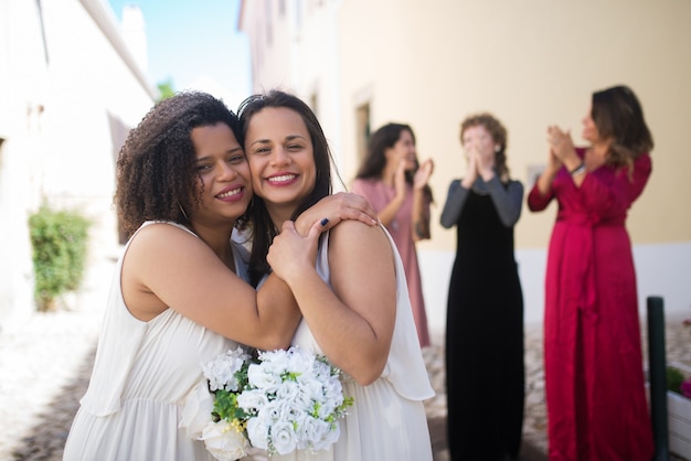Портрет милых улыбающихся невест. Две молодые женщины обнимаются. Гости-женщины смеются и аплодируют