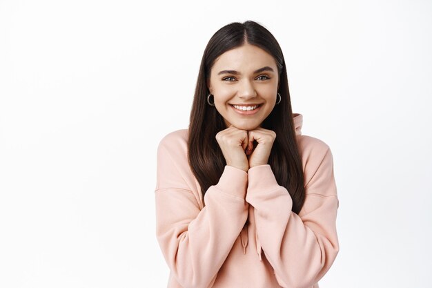 Portrait of cute smiling brunette girl, lean on hands and watching something interest, staring amazed at front, standing entertained at white wall