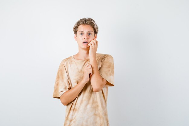 Портрет симпатичного мальчика-подростка, держащего руку за щеку в футболке и выглядящего испуганным, вид спереди