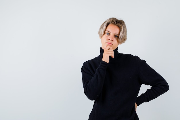 Portrait of cute teen boy keeping hand on chin in turtleneck sweater and looking thoughtful front view