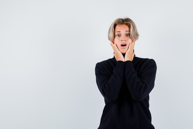 Portrait of cute teen boy keeping hands on cheeks in turtleneck sweater and looking anxious front view