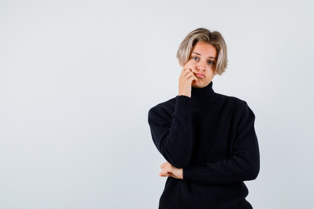 Portrait of cute teen boy leaning cheek on hand in turtleneck sweater and looking sad front view