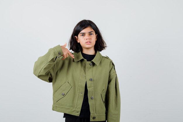 Portrait of cute teen girl pointing at herself finger pistol in army green jacket and looking upset front view