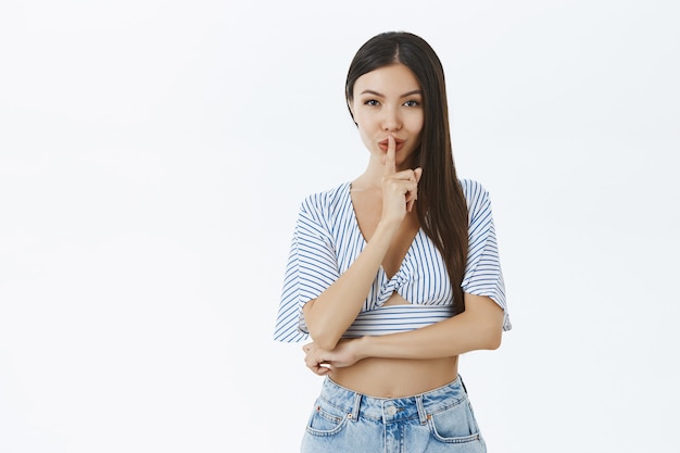 Portrait of cute and tender feminine asian girlfriend in stylish cropped blouse showing shush sign