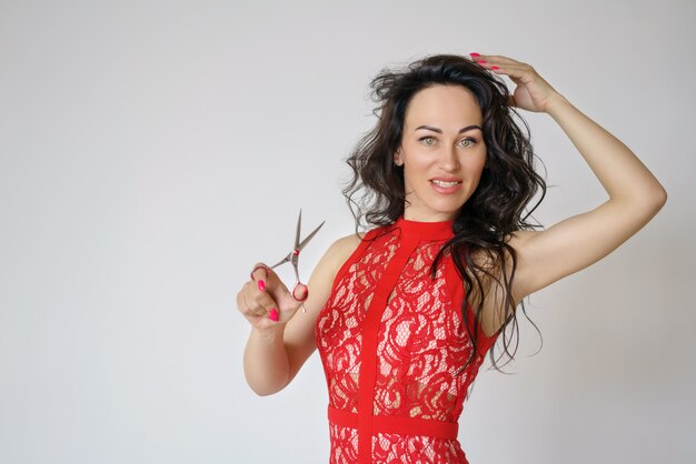 Portrait of a cute woman in a red dress with long hair holding a pair of scissors in her hand on a light