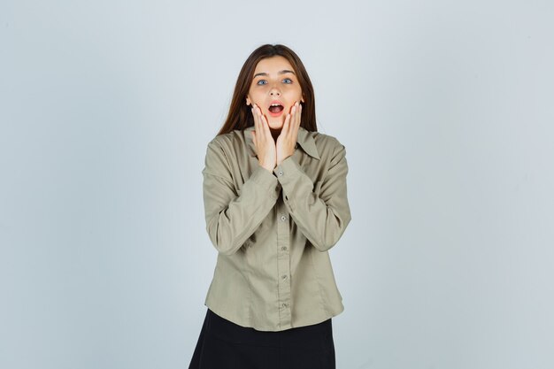 Portrait of cute young female holding hands on cheeks in shirt, skirt and looking surprised front view