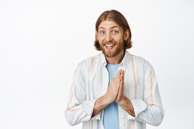 Portrait of cute young man, bearded guy thanking you, holding hands in pray and smiling, asking for favour, beggig for smth, standing against white background.