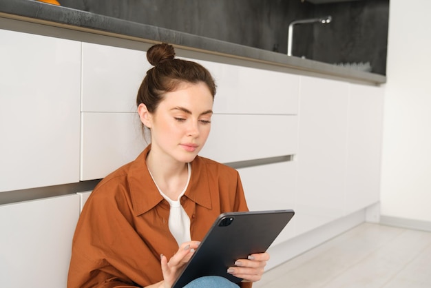 Foto gratuita ritratto di una carina giovane donna freelance che lavora da casa seduta sul pavimento della cucina con digitale