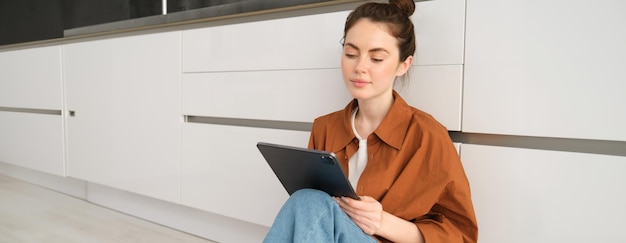 Free photo portrait of cute young woman freelancer working from home sitting on kitchen floor with digital