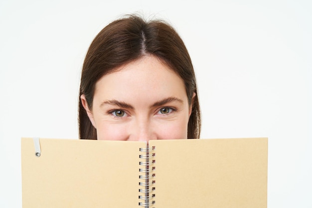 Free photo portrait of cute young woman hides her face behind planner holds notebook against her face and