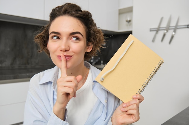 Free photo portrait of cute young woman holds planner writes her secrets in diary shows hush shush gesture